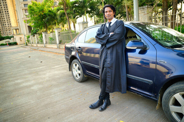 Lawyer standing next to car, A man in a graduation gown stand, With his arms crossed, smiling, The background features a fence and trees, Indian, Looking at camera, Smile Judge, Magistrate, Justice.