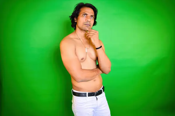 stock image Shirtless man posing thoughtfully, Men thinking pose, Looking up while think, Indian, With long hair, stands against a green background, Looks thoughtful, Hand on his chin, White pant, Pensive male.