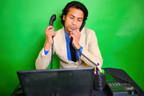 İş adamı sabit hat telefonundan konuşuyor, iki telefonla konuşuyor, bej takım elbiseli, mavi gömlekli bir adam, elinde dizüstü bilgisayarla bir masada oturuyor, yeşil bir geçmişi var, Hindistanlı, yönetici....