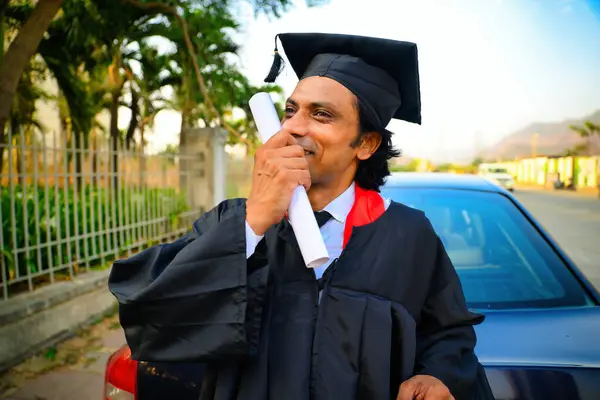 stock image Man celebrating graduation outdoors, A man wearing a graduation cap and gown, Holding diploma and smiling. He is standing outdoors near a parked car, Indian, Looking away, kissing to degree, 30s guy.