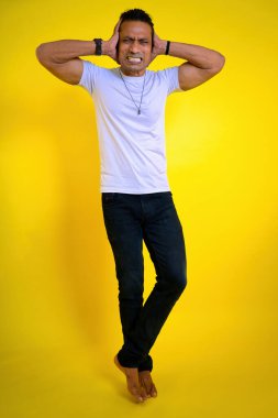 Öfkeli adam kulaklarını kapatıyor çapraz bacaklı adam sarı arka planda, hayal kırıklığına uğramış yüz ifadesinde. Acı içinde bağırıyordu. Aklı başında değil, gürültü bunalımı, Hintli, kaslı vücut, 30 yaşında.