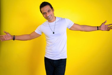 Neşeli Adam Kollarını Açarak Karşılıyor. Beyaz tişörtlü, kollarını açmış gülümsüyor. Sarı arka plana karşı. Genç mutlu çocuk ayakta duruyor. Sarılma hareketi gösteriliyor. Sarılma konsepti, Hint, Asyalı, 30 'lu yaşlarda.