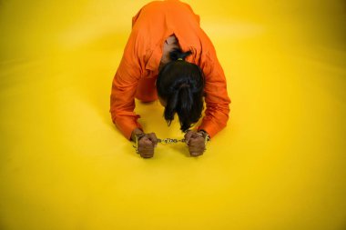 Kelepçeli mahkum yere eğiliyor turuncu tulumlu adam sarı arka planda, uzun saçlı ve at kuyruklu suçlu yardım istiyor özgürlük hapishanesi, Hintli, Asyalı, 30 'lu yaşların arkasındaki