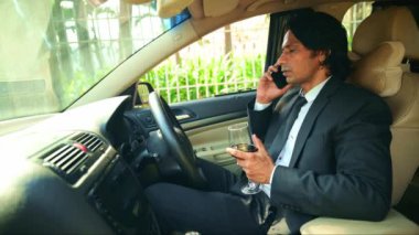 Takım elbiseli genç Hintli işadamı arabada otururken cep telefonuyla konuşuyor ve elinde bir kadeh şarap tutuyor.