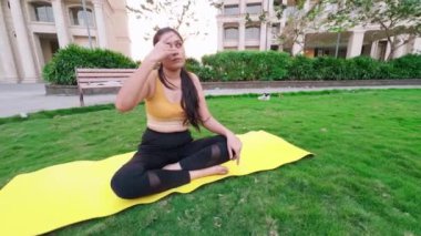 Spor kıyafetleriyle yoga yapan genç Hintli bir kız, şehir parkında meditasyon yapan bir kadın, bahçedeki çimlerin üzerinde yoga minderinde oturan uzun saçlı bir kız.   