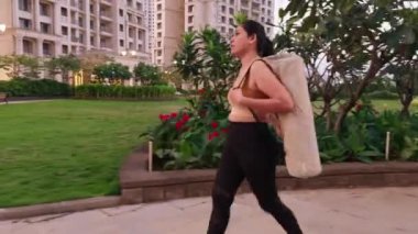 Spor kıyafetleriyle yürüyen genç bir bayan, inşaat yolunda yürüyen bir kadın, yoga minderi taşıyan güzel bir kadın, Hintli.
