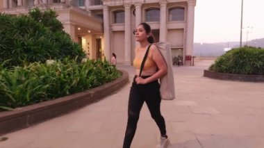 Konut kompleksinde yürüyen bir kadın, patikada yürüyen genç bir bayan, yoga minderiyle yürüyen bir kız sporcu.  