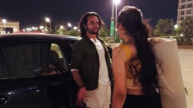 Arka planda yüksek binalar olan Hintli bir çift gece araba yanında tartışıyor.