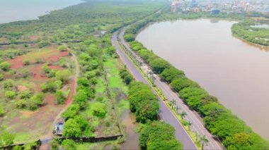 Göl kenarındaki bir otoyolun hava görüntüsü, insansız hava aracı Mumbai şehir manzarası, çevresindeki yemyeşil ve şehir binaları, arka planda yüksek kuleler, göle paralel giden trafik, Hindistan