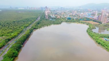 Göl kenarındaki bir otoyolun hava görüntüsü, insansız hava aracı Mumbai şehir manzarası, çevresindeki yemyeşil ve şehir binaları, arka planda yüksek kuleler, göle paralel giden trafik, Hindistan