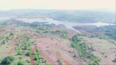 Yeşilliklerle çevrili küçük bir kasaba, kırmızı topraklı tarım arazisi, arka planda nehir gölü, Uzak tepeler, Hindistan