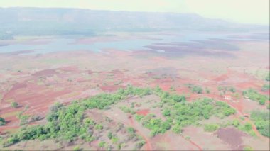 Yeşilliklerle çevrili küçük bir kasaba, kırmızı topraklı tarım arazisi, arka planda nehir gölü, Uzak tepeler, Hindistan