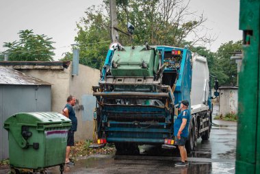  Çöp toplama işlemi. İki kişi çöp konteynırlarını çöp kamyonuna çekiyor ve çöplerin yüklenmesini denetliyor..
