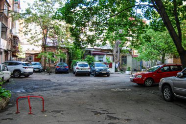 Park halindeki arabaları ve binaları olan bir konut avlusu. Birkaç park etmiş aracın çevresi alçak binalarla çevrili.. 