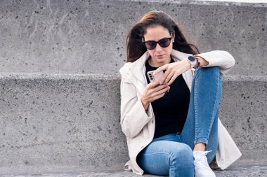 Kafkasyalı esmer kız büyük beton bir bankta oturmuş akıllı telefonunu kontrol ediyor..