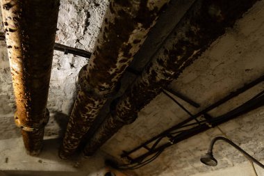 Ağır pas ve oksitlenmiş yüzeyli eski borular terk edilmiş bir madenin yeraltı sanayi koridorunda, düşük ışık ve kumlu detaylar..