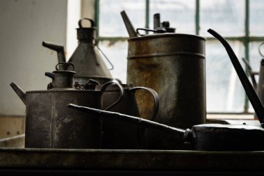 Eski metal yağ bidonları ve sürahiler endüstriyel bir atölyede, pencere ışığıyla aydınlatılmış, pas ve patinajı gösteren.