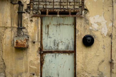 Eski paslı metal kapı, soyulmuş boya ve yıpranmış endüstriyel bir duvarın üzerinde paslanmış çerçeve ve elektrik kutusu..