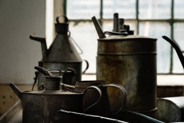 Eski metal yağ bidonları ve sürahiler endüstriyel bir atölyede, pencere ışığıyla aydınlatılmış, pas ve patinajı gösteren.
