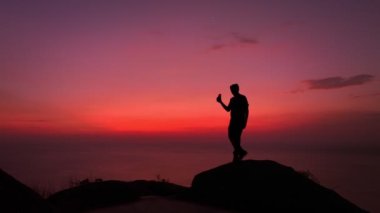 Kızıl gökyüzü ve deniz manzaralı bir sahneye karşı bir adam durur ve selfie çeker gökyüzünde ve deniz manzarası sarp kayalıklarda kırmızıdır alacakaranlık kırmızı gökyüzü..