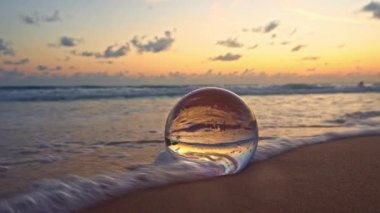 Akşamları parlak renklerde güzel gökyüzü kumsaldaki kristal kürenin içinde. Altın plajdaki kristal küre, güzel ve alışılmadık seyahat fikirleri için dalga 4K Videolar tarafından yuvarlanıyor..