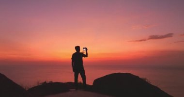 Uçurumun kenarında duran ve kızıl gökyüzüne karşı selfie çeken bir adam..