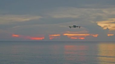 Hobi olarak ve rahatlamak için deniz resimlerini çekmek için bir dron uçurmak. Altın plajın, derin mavi okyanus suyuyla ve köpüklü dalgalarla buluşmasının güzel günbatımında kumsalda uçan dron görüntüsü..