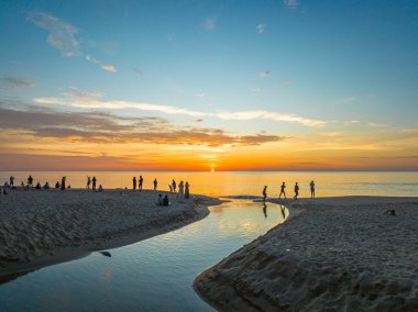 Kumsaldaki dolambaçlı kanalın hava görüntüsü. Turistler kumlu bir sahildeki küçük bir kanalın yanında batan güneşi seyrediyorlar. Kanaldaki güzel gökyüzü yansıması..