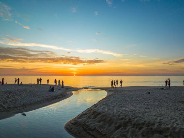 Kumsaldaki dolambaçlı kanalın hava görüntüsü. Turistler kumlu bir sahildeki küçük bir kanalın yanında batan güneşi seyrediyorlar. Kanaldaki güzel gökyüzü yansıması..