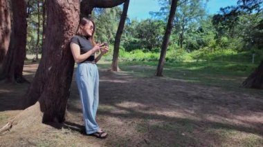  Bir kadın büyük bir çam ağacının gölgesinde cep telefonu kullanarak rahatça ayakta duruyor.