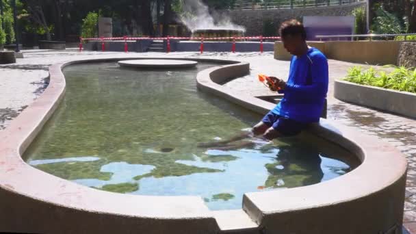 一名男子在温泉浴场坐在温泉池的座位上 坐在温泉池中观看手机 游客们把脚浸在温泉池里放松一下 — 图库视频影像