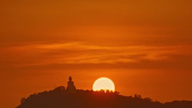 Dağdaki Buda heykelinin arkasında yuvarlak sarı bir güneş batıyor..