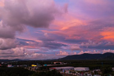 Phuket kırsalında canlı bir günbatımını yakalayan nefes kesici bir hava manzarası. Gökyüzü sarı, turuncu, pembe ve mor renkli zengin renklerle parlarken dramatik bulutlar şehrin üzerinde süzülüyor.