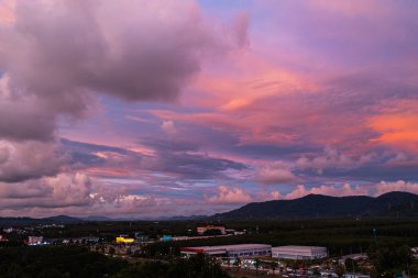 Phuket kırsalında canlı bir günbatımını yakalayan nefes kesici bir hava manzarası. Gökyüzü sarı, turuncu, pembe ve mor renkli zengin renklerle parlarken dramatik bulutlar şehrin üzerinde süzülüyor.