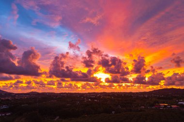 Phuket kırsalında canlı bir günbatımını yakalayan nefes kesici bir hava manzarası. Gökyüzü sarı, turuncu, pembe ve mor renkli zengin renklerle parlarken dramatik bulutlar şehrin üzerinde süzülüyor.