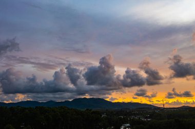 Phuket kırsalında canlı bir günbatımını yakalayan nefes kesici bir hava manzarası. Gökyüzü sarı, turuncu, pembe ve mor renkli zengin renklerle parlarken dramatik bulutlar şehrin üzerinde süzülüyor.