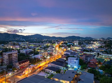 Alacakaranlıktaki Phuket şehrinin büyüleyici hava manzarası sokakları aydınlatan canlı şehir ışıklarıyla. Gökyüzünün yumuşak renkleri uzak ufukla kusursuz bir şekilde harmanlanarak, dinginlik yaratır.