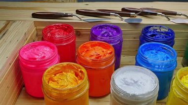  Açık akrilik boya kavanozlarından oluşan renkli bir düzenleme. Sıcak kırmızı ve portakallardan serin mavi ve yeşilliklere kadar canlı renkler içeriyor. Arkadaki tahta kutu ve palet bıçakları..