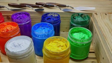  Açık akrilik boya kavanozlarından oluşan renkli bir düzenleme. Sıcak kırmızı ve portakallardan serin mavi ve yeşilliklere kadar canlı renkler içeriyor. Arkadaki tahta kutu ve palet bıçakları..