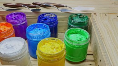  Açık akrilik boya kavanozlarından oluşan renkli bir düzenleme. Sıcak kırmızı ve portakallardan serin mavi ve yeşilliklere kadar canlı renkler içeriyor. Arkadaki tahta kutu ve palet bıçakları..