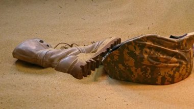 Terk edilmiş askeri teçhizat, yıpranmış bir bot, kask ve parçalanmış koruyucu gözlük, kısmen çöl kumuna gömülmüş, çatışmanın acı gerçeklerini simgeliyor..