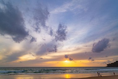Suya yansıyan yumuşak altın ışıkla okyanusun üzerinde baş döndürücü bir günbatımı, kıyıda yürüyen insanların siluetlerini içeriyor. Su küçük bir dereden denize akar..