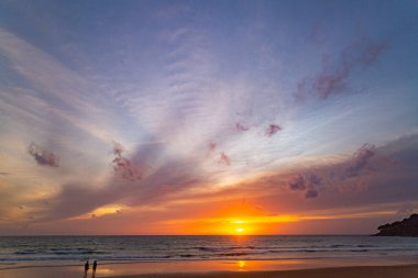 Suya yansıyan yumuşak altın ışıkla okyanusun üzerinde baş döndürücü bir günbatımı, kıyıda yürüyen insanların siluetlerini içeriyor. Su küçük bir dereden denize akar..