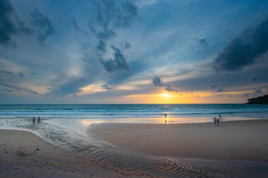 Suya yansıyan yumuşak altın ışıkla okyanusun üzerinde çarpıcı bir günbatımı, kıyı boyunca yürüyen insanların siluetlerini içeriyor. Su küçük bir dereden denize akıyor.