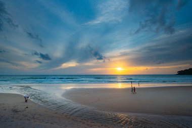Suya yansıyan yumuşak altın ışıkla okyanusun üzerinde çarpıcı bir günbatımı, kıyı boyunca yürüyen insanların siluetlerini içeriyor. Su küçük bir dereden denize akıyor.