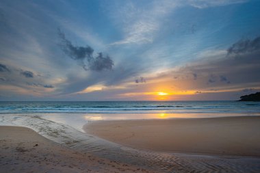 Suya yansıyan yumuşak altın ışıkla okyanusun üzerinde çarpıcı bir günbatımı, kıyı boyunca yürüyen insanların siluetlerini içeriyor. Su küçük bir dereden denize akıyor.