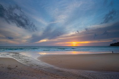 Suya yansıyan yumuşak altın ışıkla okyanusun üzerinde çarpıcı bir günbatımı, kıyı boyunca yürüyen insanların siluetlerini içeriyor. Su küçük bir dereden denize akıyor.