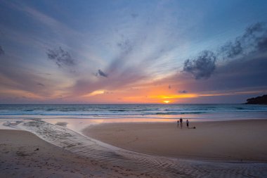 Suya yansıyan yumuşak altın ışıkla okyanusun üzerinde çarpıcı bir günbatımı, kıyı boyunca yürüyen insanların siluetlerini içeriyor. Su küçük bir dereden denize akıyor.