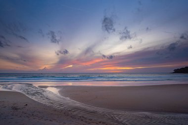 Alacakaranlıkta sakin bir sahil sahnesi, canlı bir günbatımı sergiliyor sakin sulara ve ıslak kuma yansıyan renkli bulutlarla. Karon plajında gün batımında muhteşem bir gökyüzü. tatlı gökyüzü arkaplanı