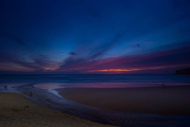 Alacakaranlıkta sakin bir sahil sahnesi, canlı bir günbatımı sergiliyor sakin sulara ve ıslak kuma yansıyan renkli bulutlarla. Karon plajında gün batımında muhteşem bir gökyüzü. tatlı gökyüzü arkaplanı